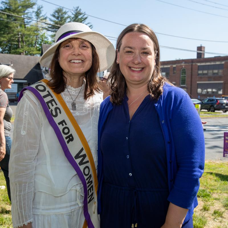 Two women stand outside wearing purple sashes and smiling.