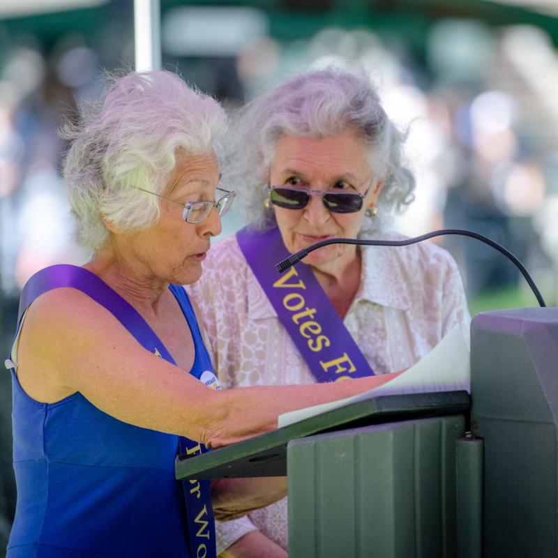 Two women wearing purple sash stand outside speaking at lectern.