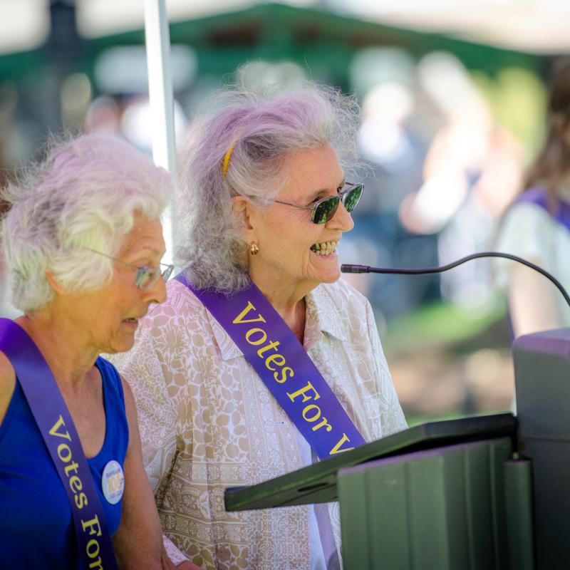 Two women stand at lectern speaking, wearing purple sashes.