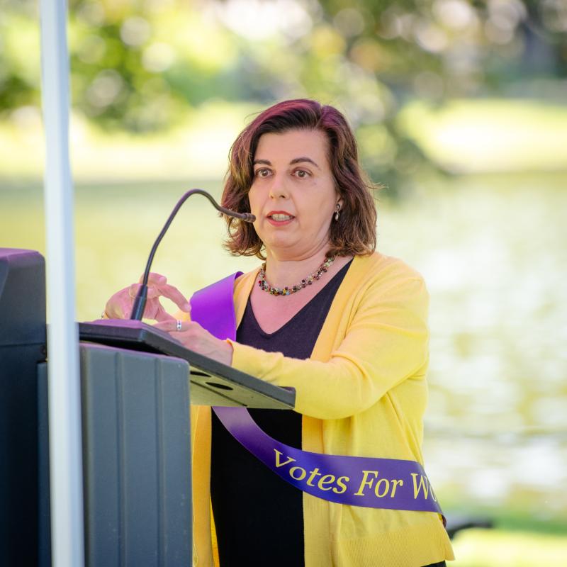 Woman wearing purple sash stands outside speaking at lectern.