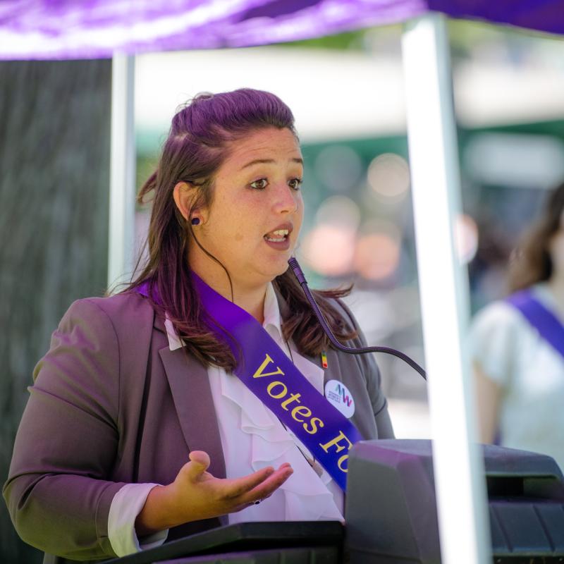 Woman wearing purple sash stands outside speaking at lectern.
