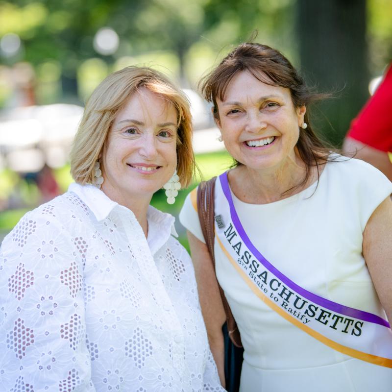 Two women wearing purple sashes stand outside smiling.