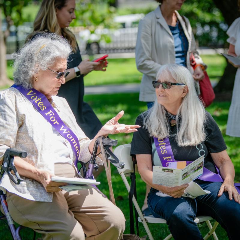 Two women sit outside wearing purple sashes.