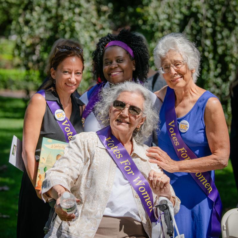 Four women in purple sashes stand and sit outside smiling.