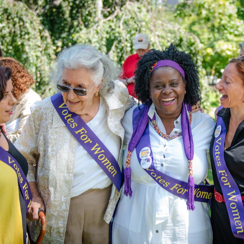 Four women stand outside laughing wearing purple sashes.