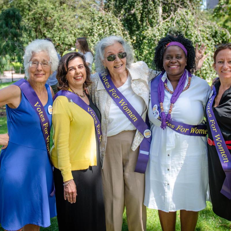 Five women stand outside wearing purple sashes.