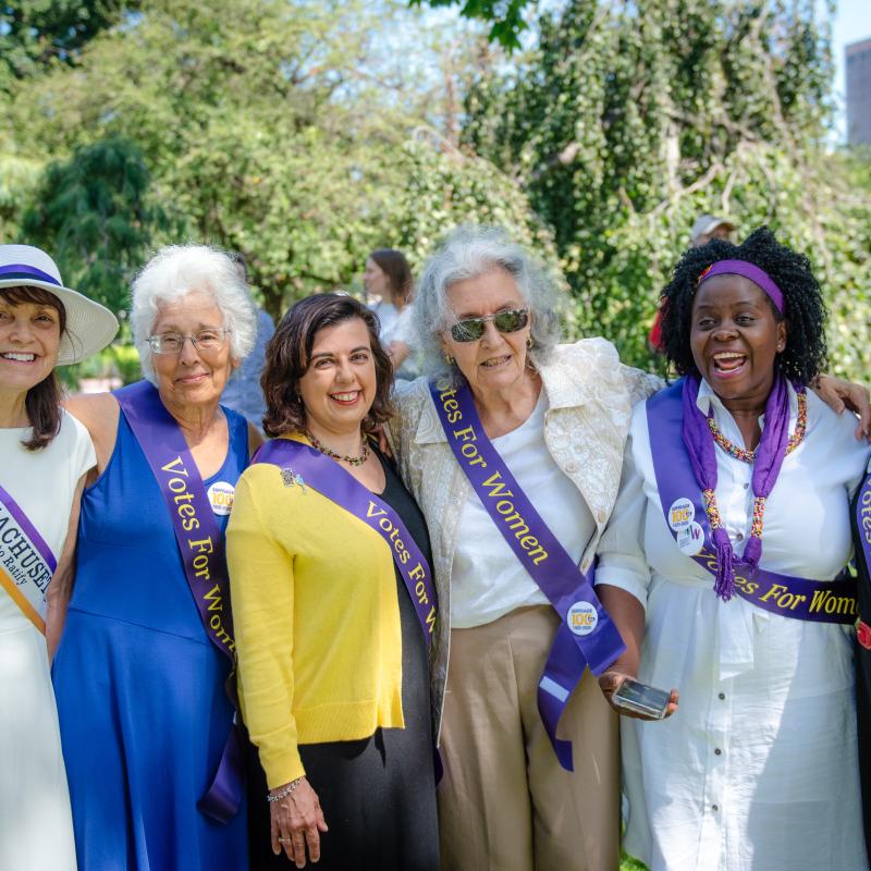 Six women stand outside wearing purple sashes.