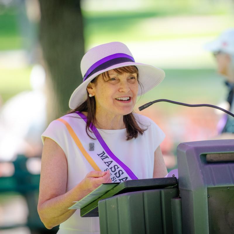 Fredie Kay wears purple sash and speaks at lectern.