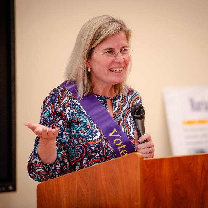 Woman wearing purple sash speaking at podium.