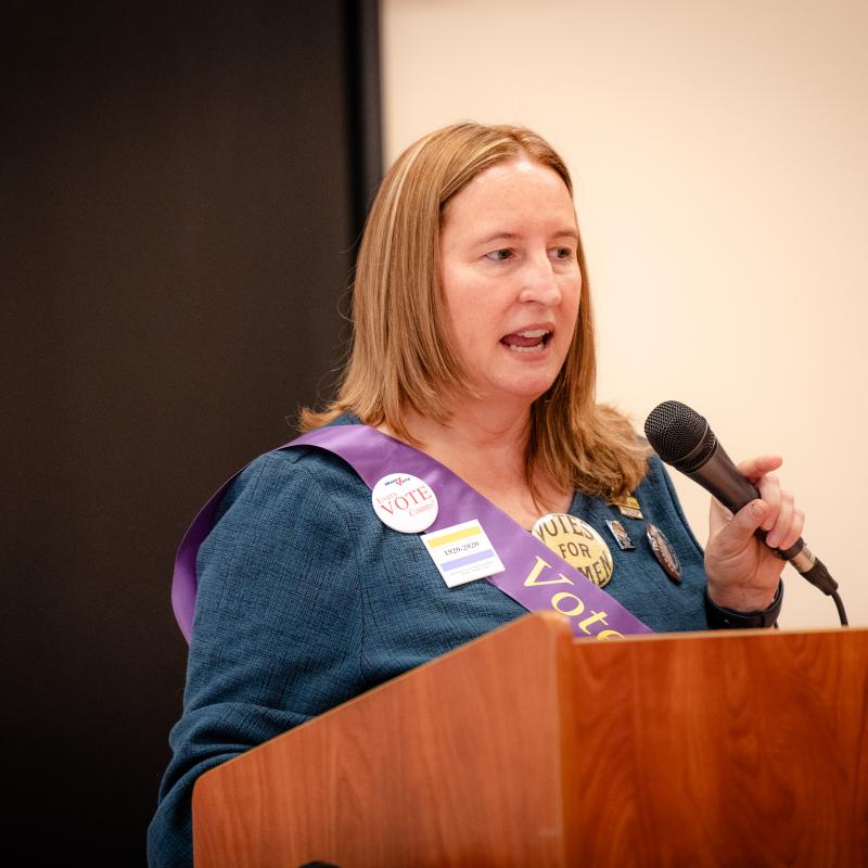 Woman wearing purple sash stands at podium holding microphone.