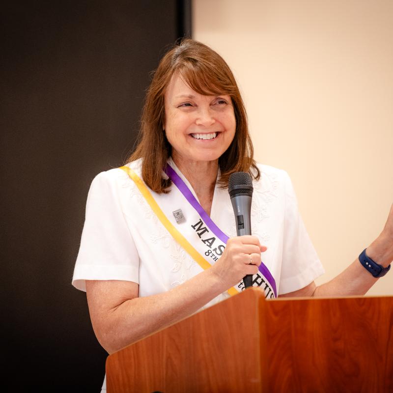 Woman wearing purple sash stands at podium holding microphone.
