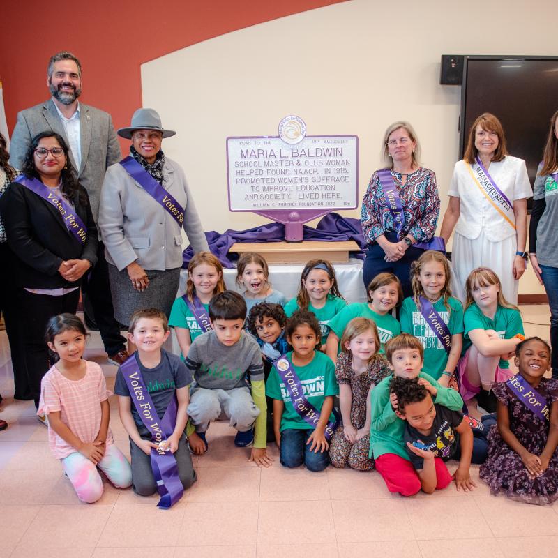 Group of adults and children stand and sit around purple and white sign.