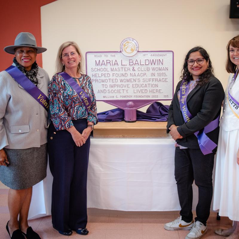 Four women wearing purple sashes stand in front of purple and white sign.