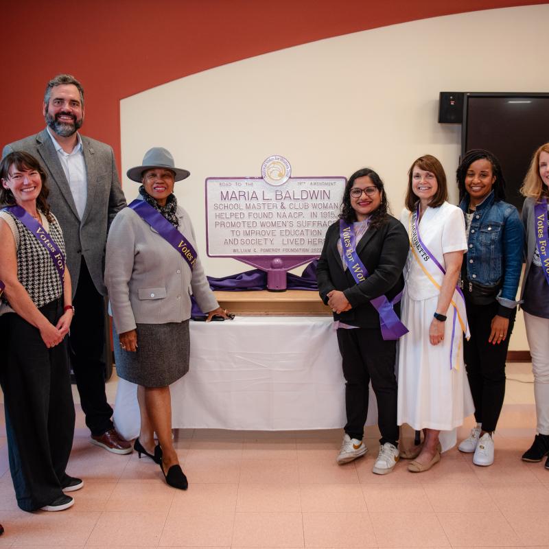 Nine adults stand next to purple and white sign.
