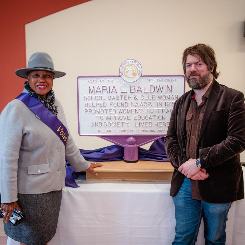 Woman and man stand next to purple and white sign.