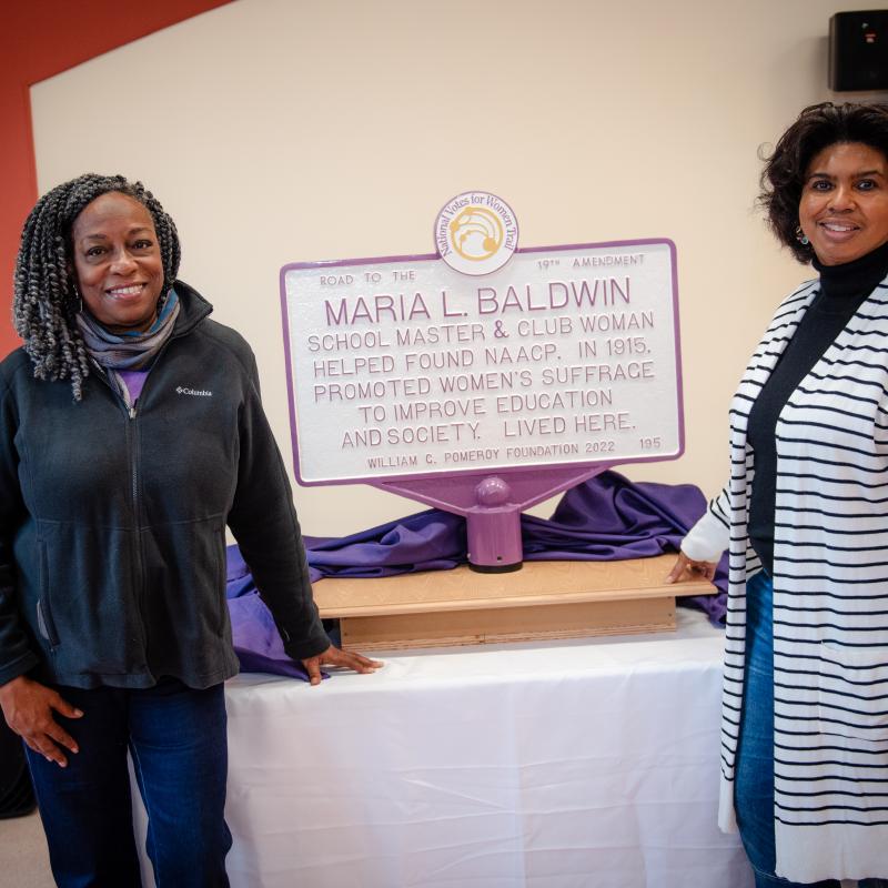 Two women stand next to Maria L. Baldwin purple and white sign.
