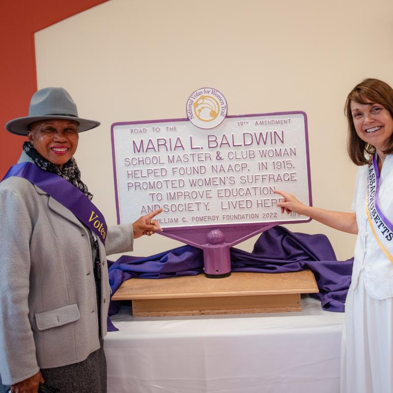 Two women stand next to Maria L. Baldwin purple and white sign.