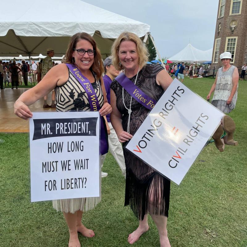 Two women stand outside wearing purple sashes and holding signs.