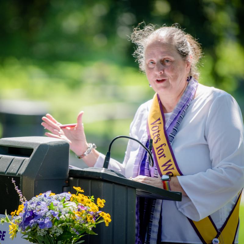 Woman wearing purple sash speaking at podium.