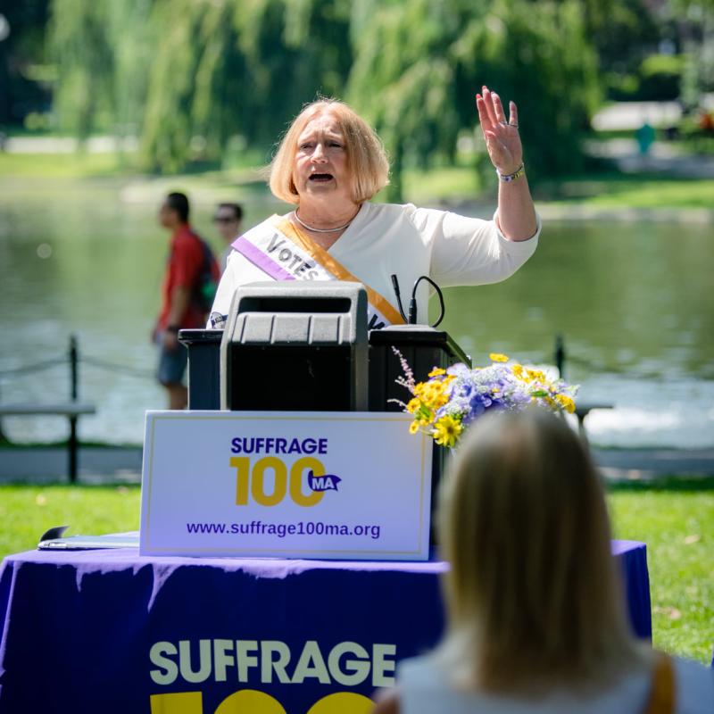 Woman wearing purple sash stands outside speaking at lectern.