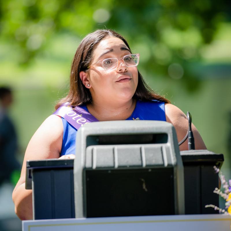 Woman wearing purple sash stands outside speaking at lectern.