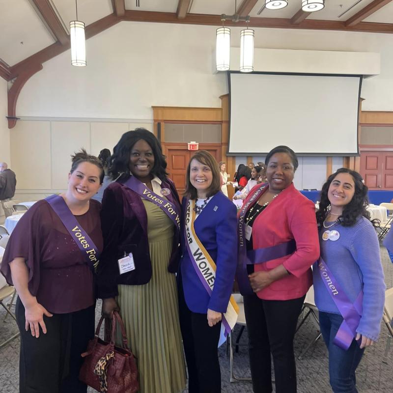 Five women wearing purple sashes stand indoors smiling.