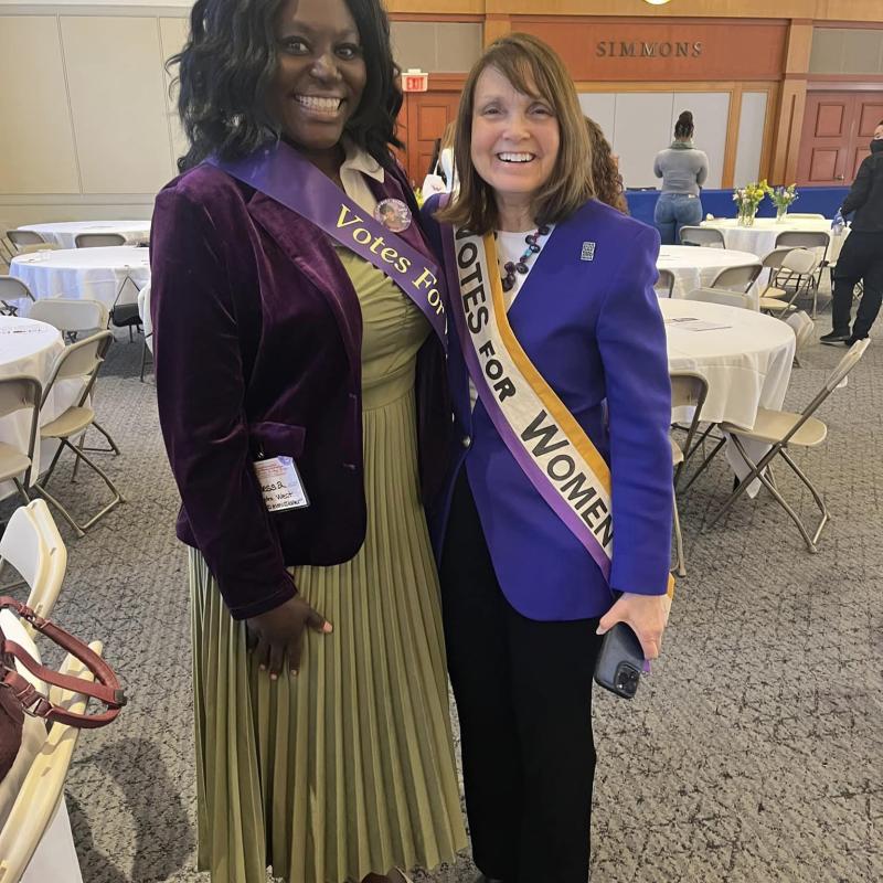 Two women wearing purple sashes stand indoors smiling.