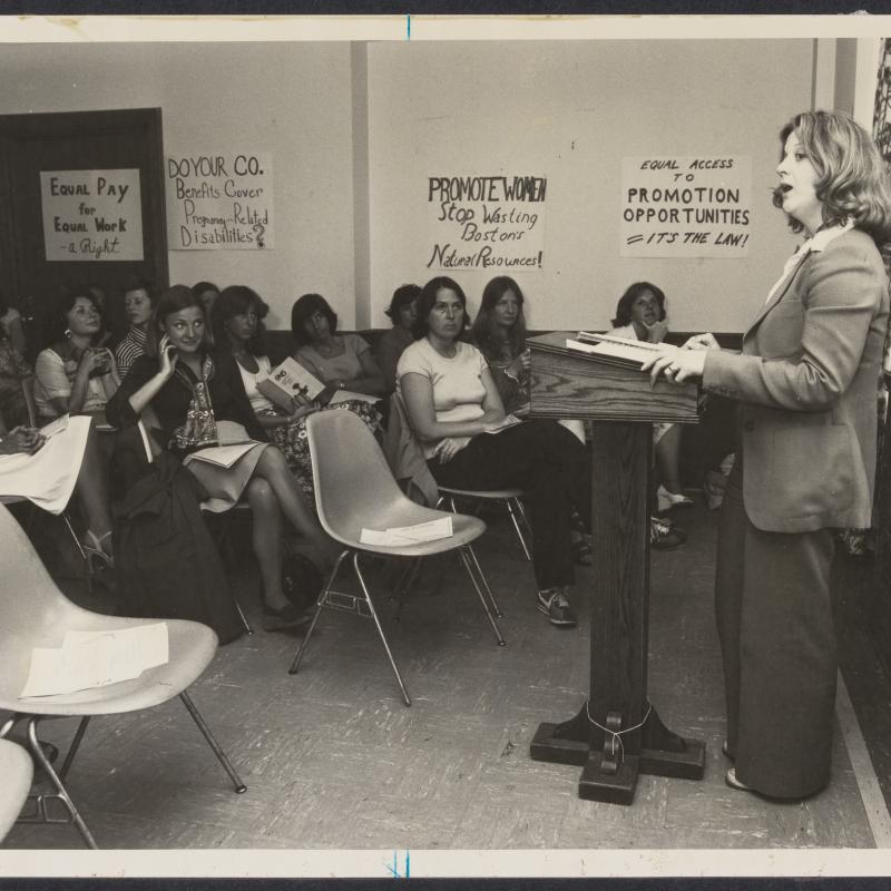 woman speaking in a meeting room in front of a group of women. Posters about equal pay and insurance companies line the walls.