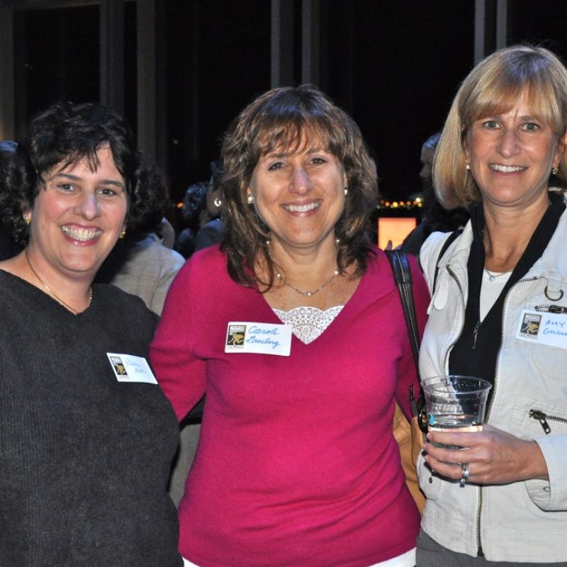 Three women stand indoors smiling.