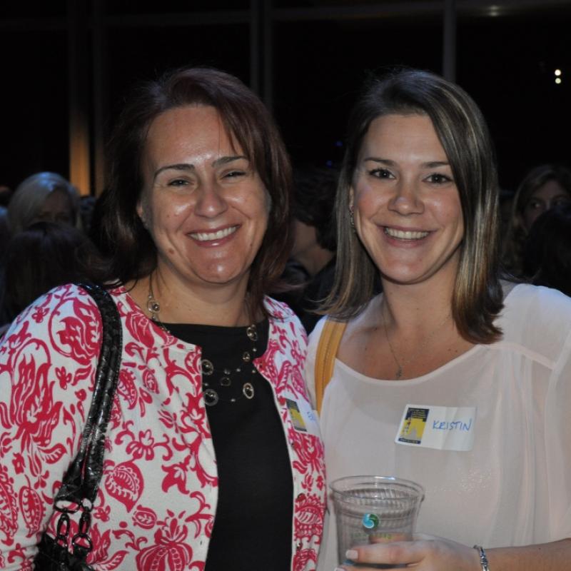 Two women stand indoors smiling.