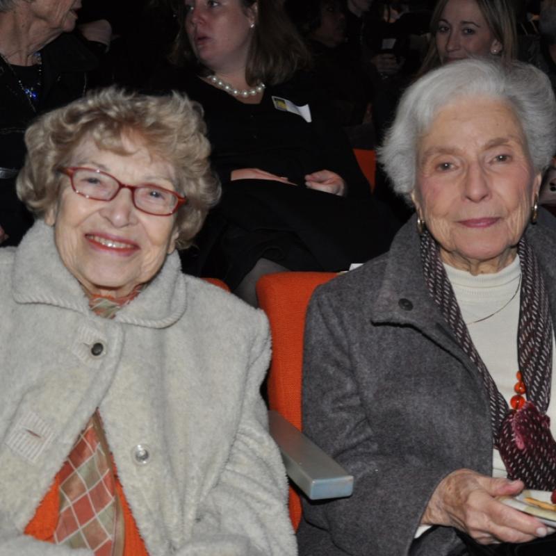 Two women sit indoors smiling.