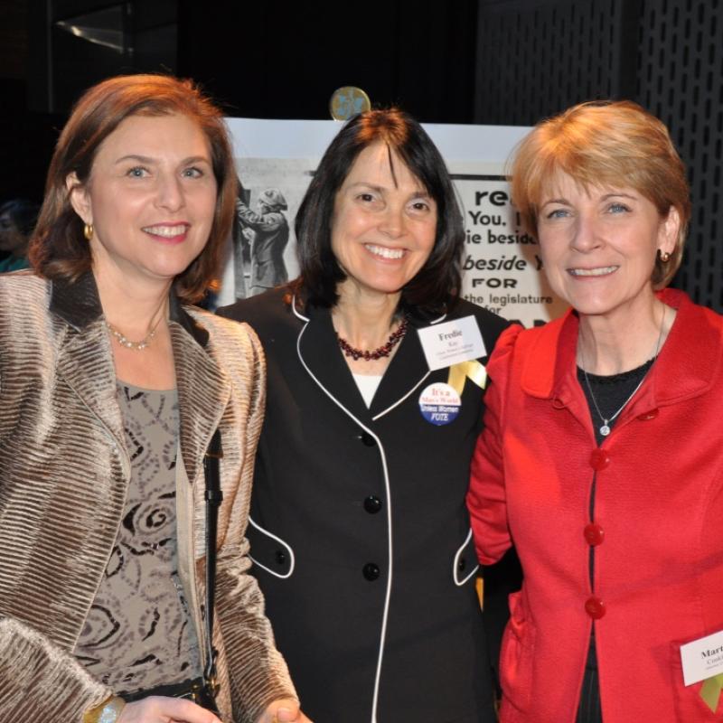 Three women stand indoors smiling.