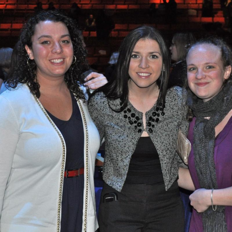 Three women stand indoors smiling.