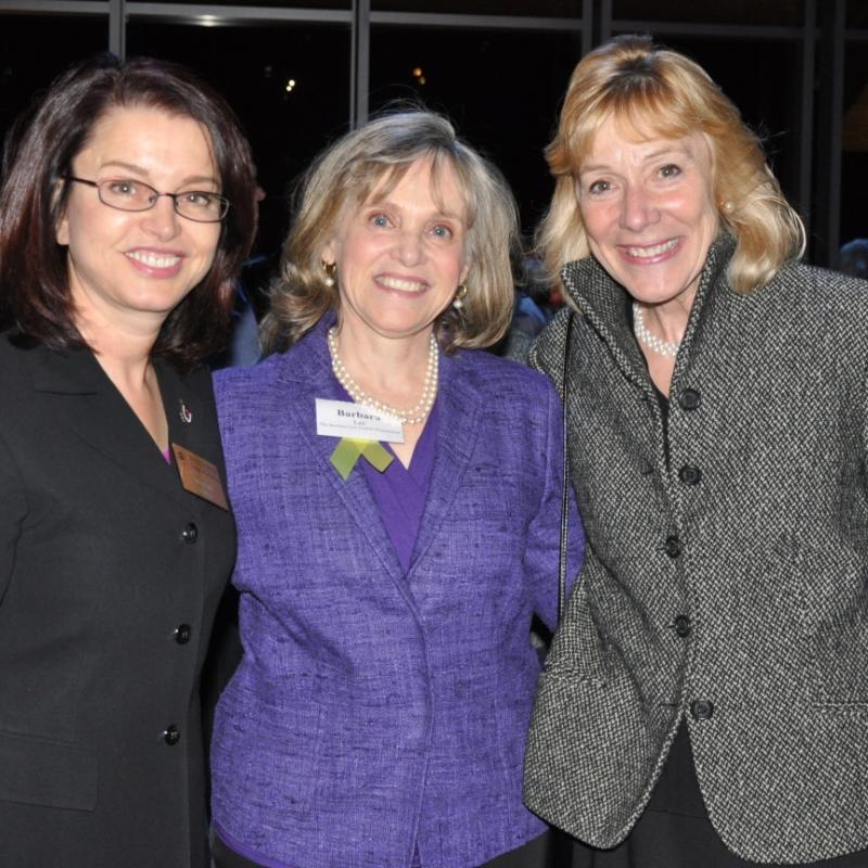 Three women stand indoors smiling.