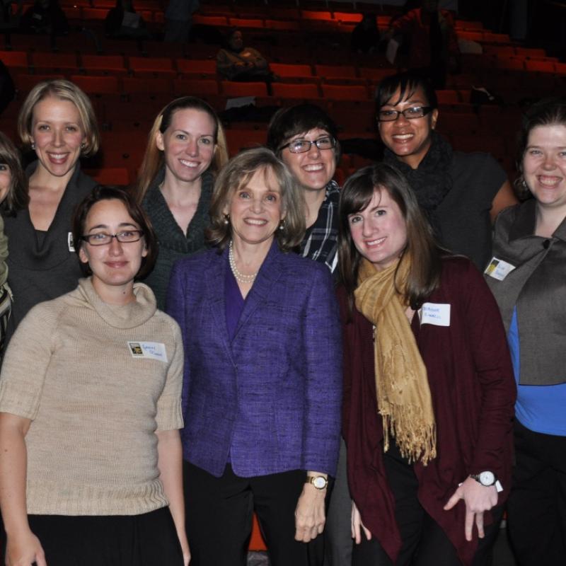 Nine women stand indoors smiling.