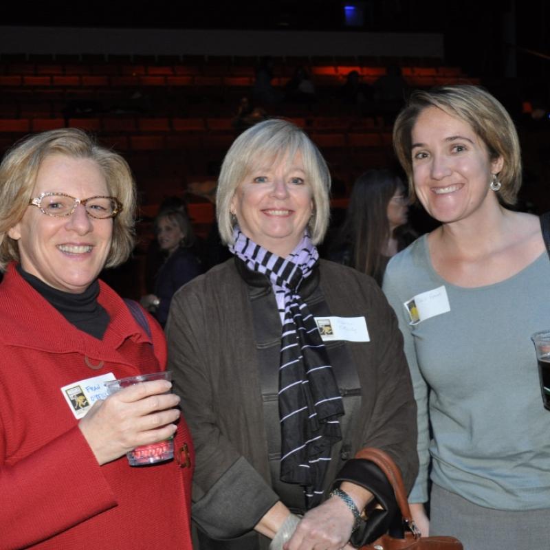 Three women stand indoors smiling.