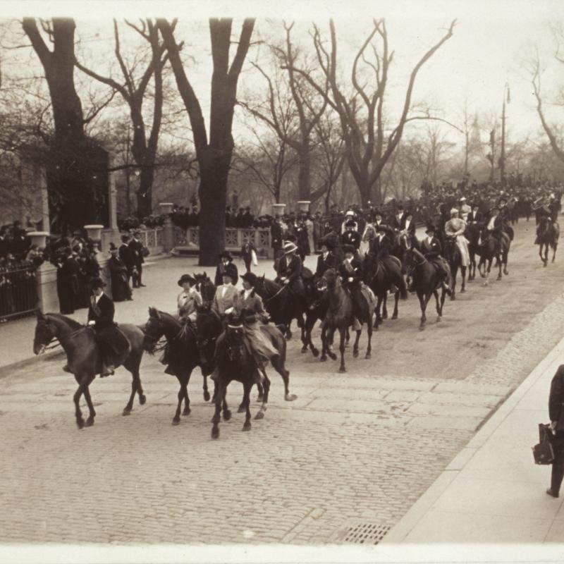  View of group of equestrian women parading down Beacon Street past the State House. The Shaw Memorial is in the background on the left. A photographer watches from the sidewalk.
