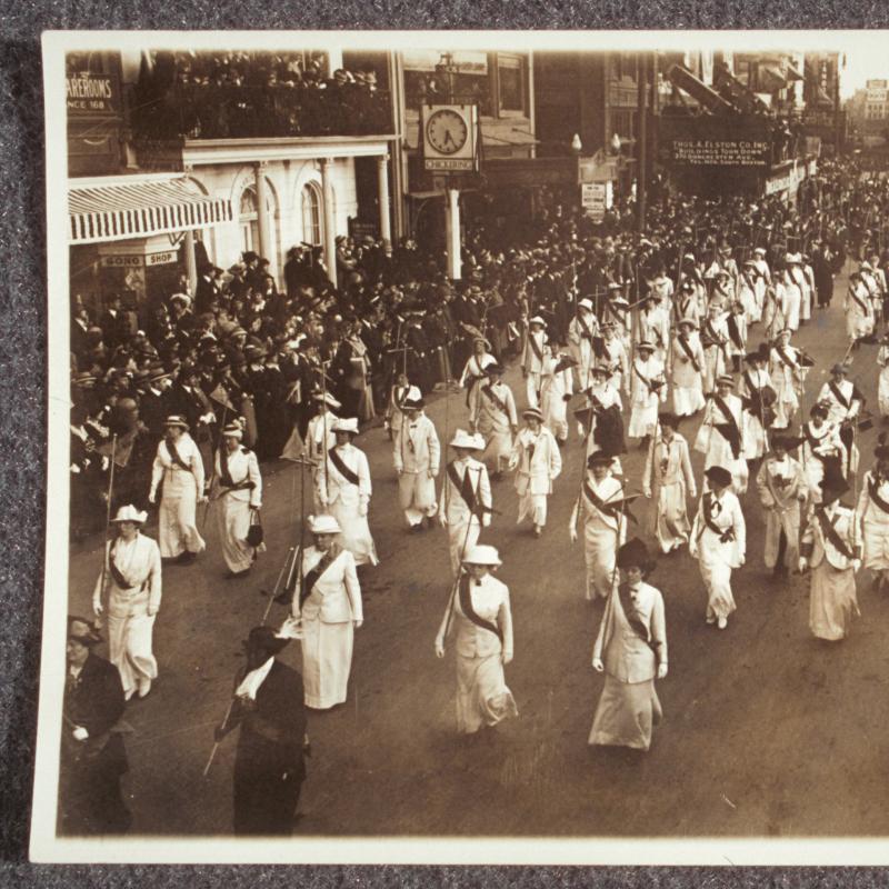 A regiment of women in uniform (white or pale clothes and a dark sash) carrying staffs and marching in formation in a suffrage parade. Crowds look on from both sidewalks.