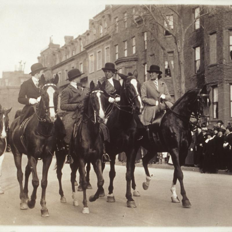 Black and white photograph of five women riding along Beacon Street on horseback in a suffrage parade. A crowd looks on from the sidewalks.