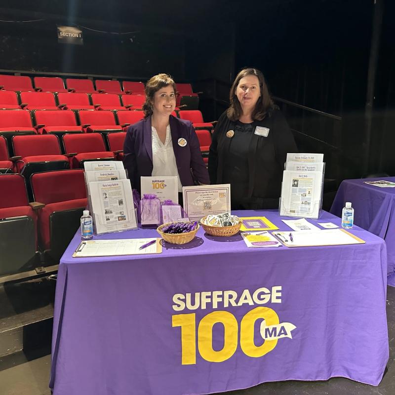 Two people stand at table with Suffrage100MA logo.