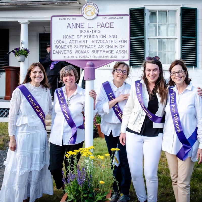 Four women wearing purple sashes stand outside in front of white and purple sign.
