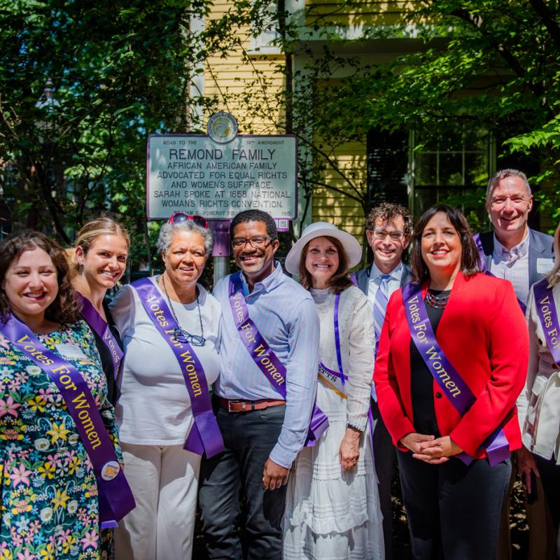 Nine people stand together wearing purple sashes and smiling.