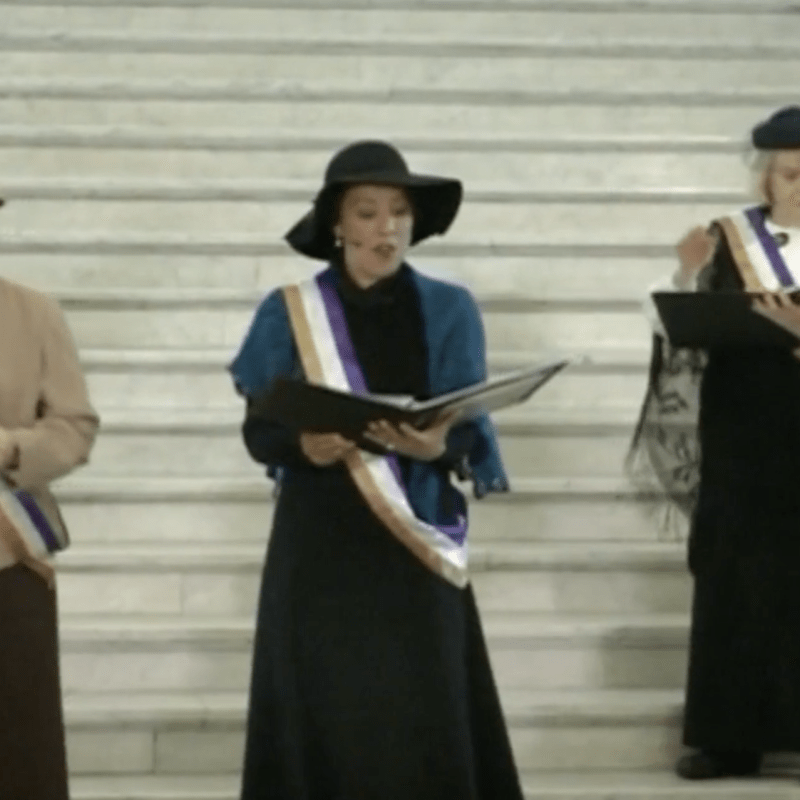 Three women stand on steps in vintage clothes, reading from programs.