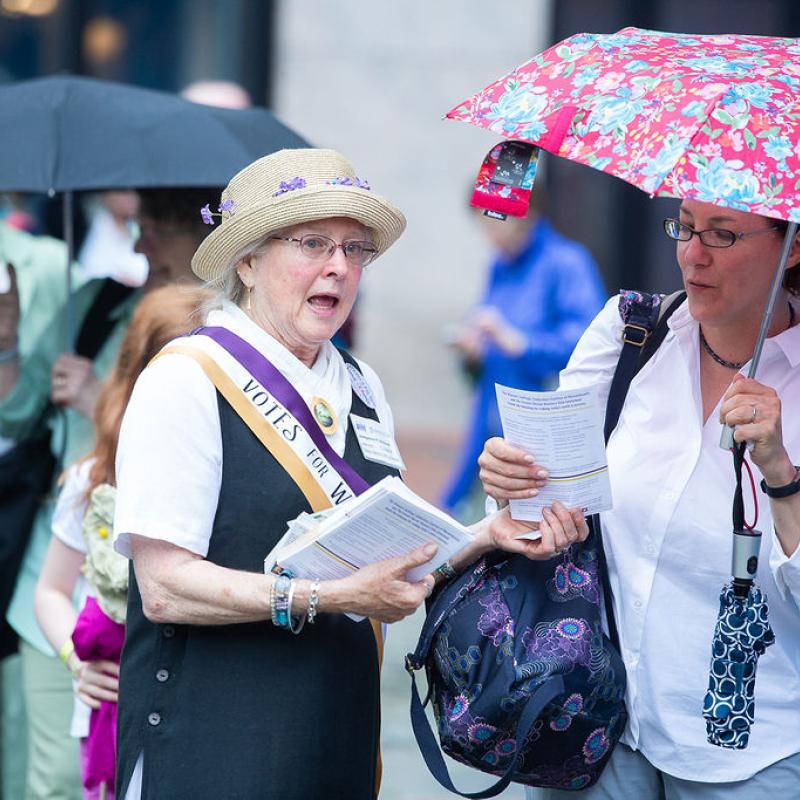 Woman wearing sash holding papers.