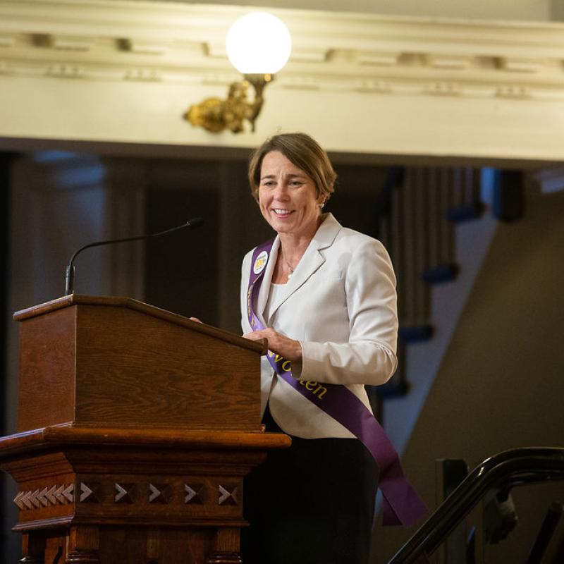 Woman wearing purple sash stands speaking at lectern.