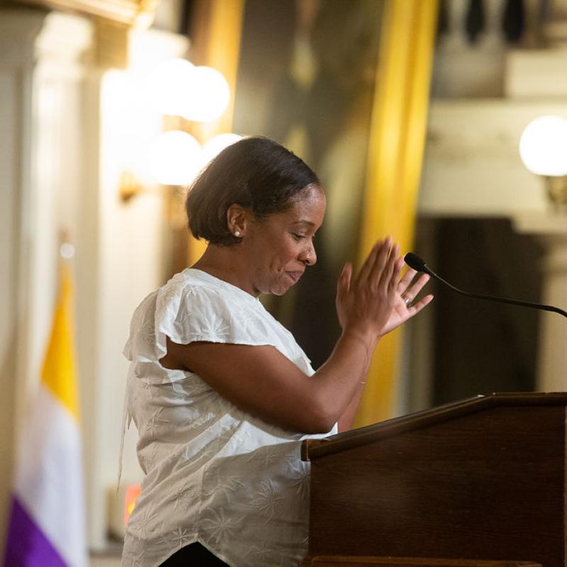 Woman wearing purple sash stands speaking at lectern.