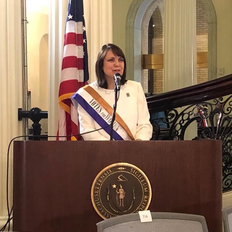 Woman wearing purple sash stands speaking at lectern.