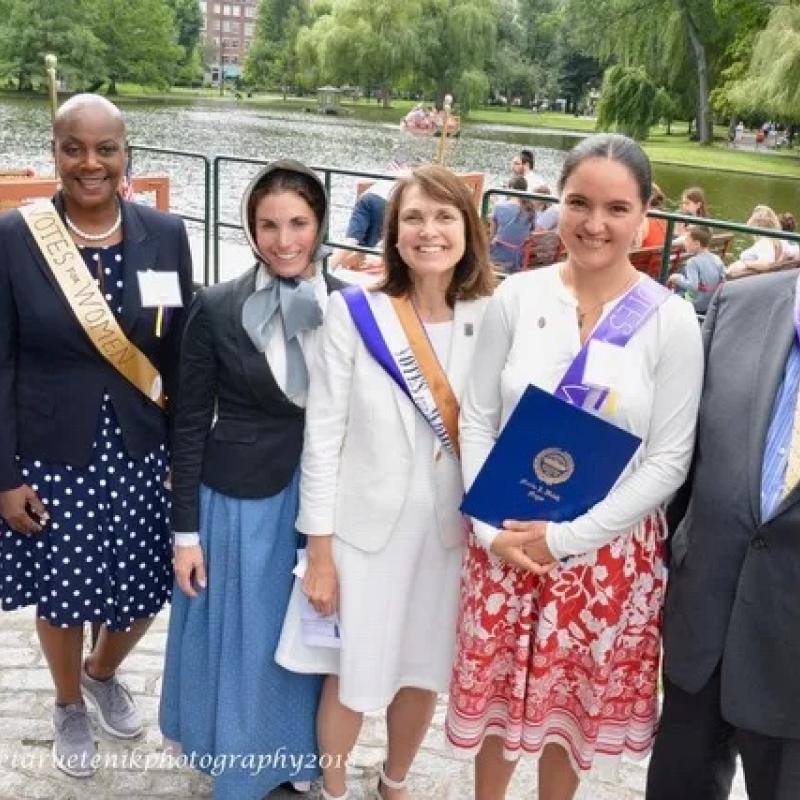 Six adults stand wearing sashes outdoors.