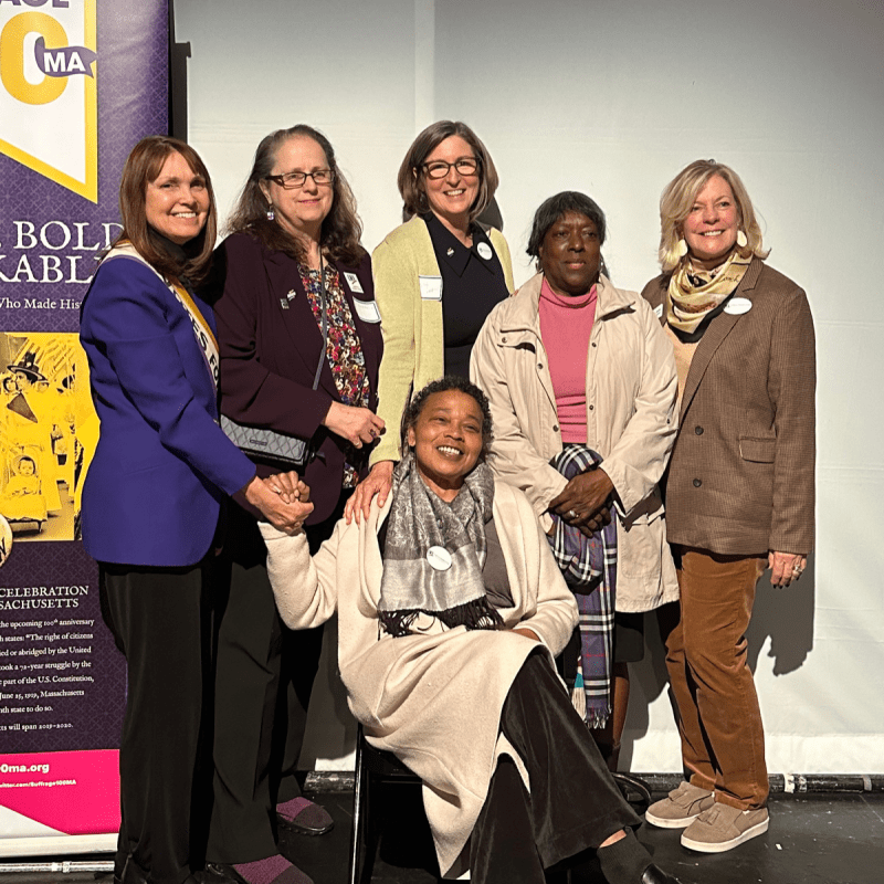 Six women stand in front of Suffrage100MA banner.