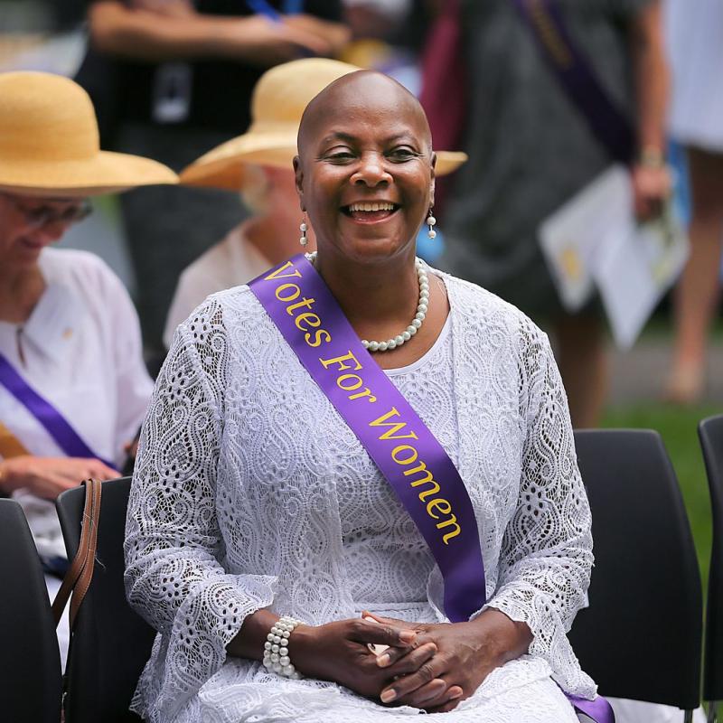 Woman wearing purple sash sits outside smiling.